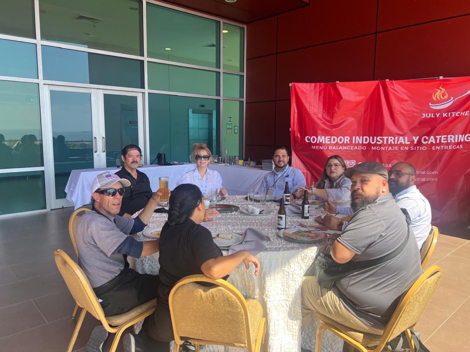 Mesa con invitados comiendo en un evento, banner de July Kitchen al fondo